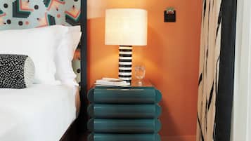 Ett hotellrum med en säng, en mönstrad sänggavel, ett turkosfärgat sidobord med en lampa, en rutig matta och en gardin.