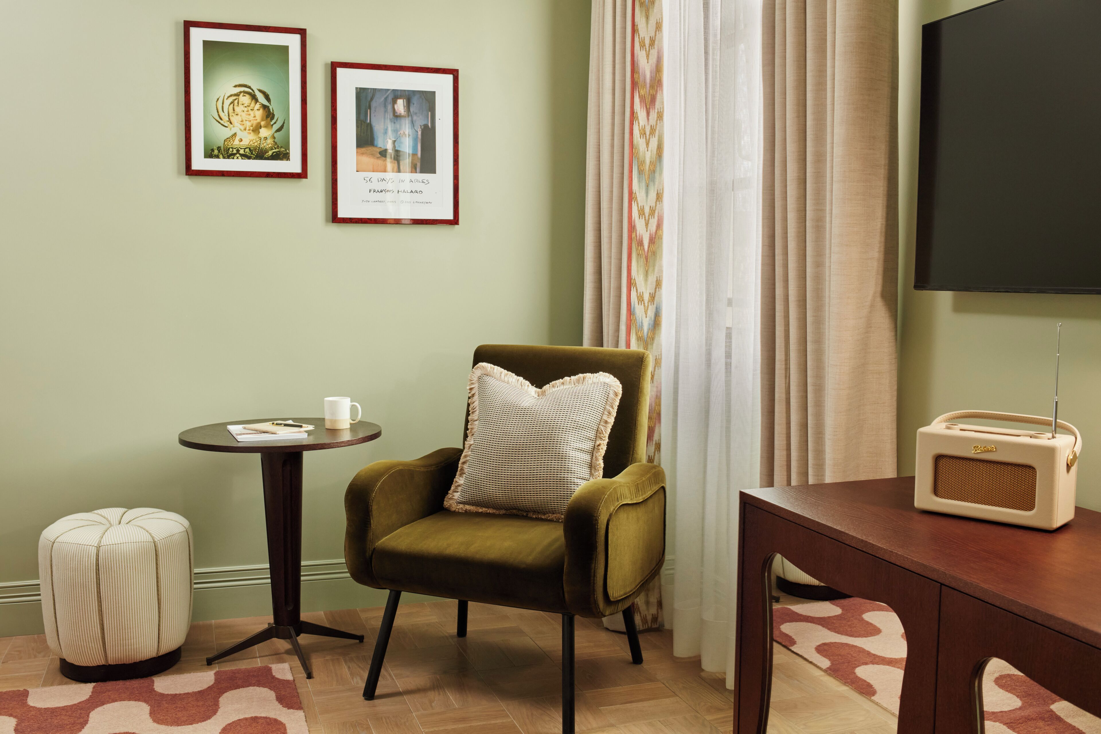 A room with a green armchair, a small round table with a cup and a book, a vintage radio, and two framed pictures on the wall.