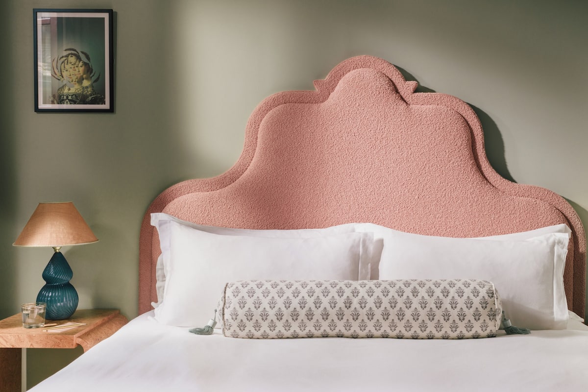 A neatly made bed with a pink textured headboard, white pillows, and a patterned throw pillow. A blue lamp and a small glass on a wooden nightstand are visible.