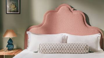 A neatly made bed with a pink textured headboard, white pillows, and a patterned pillow. A blue lamp and a glass of water are on a wooden nightstand beside the bed.