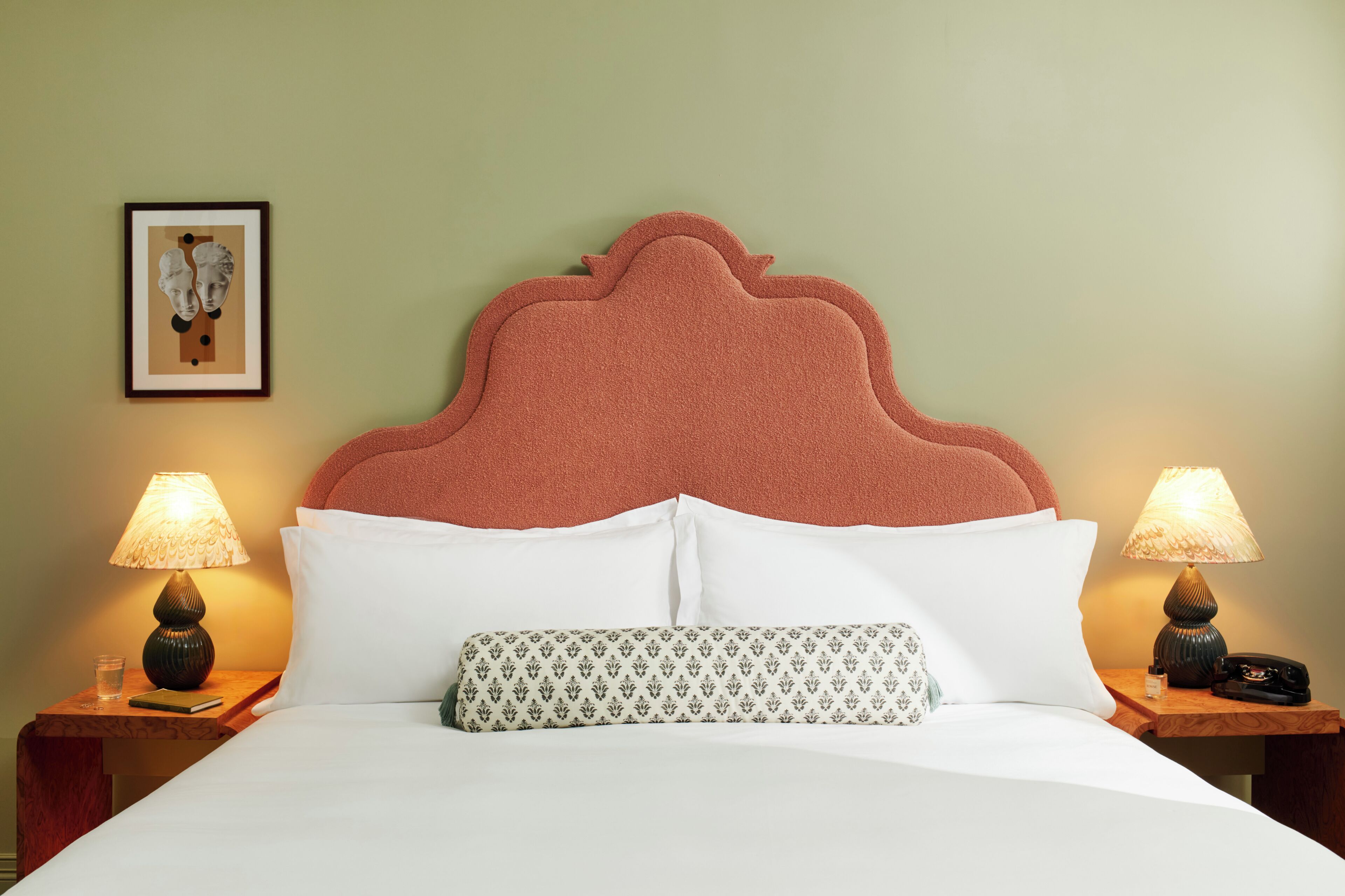 A neatly made bed with white linens, a patterned pillow, and a headboard with a scalloped design. There are two bedside tables with lamps and a framed picture on the wall.