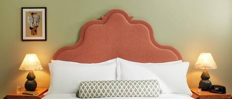 A neatly made bed with white linens, a patterned pillow, and a headboard with a scalloped design. There are two bedside tables with lamps and a framed picture on the wall.