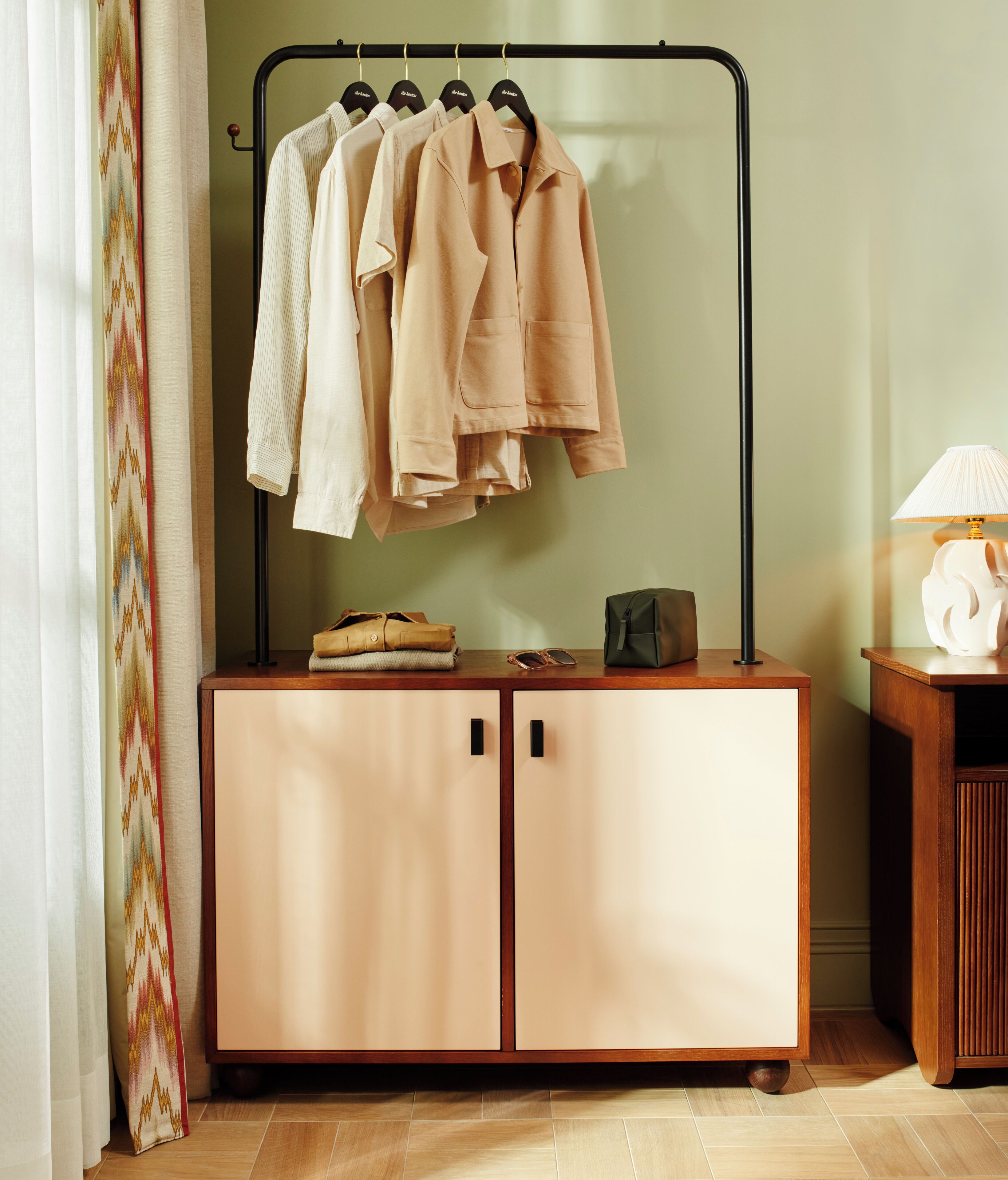 A wardrobe with hanging clothes, a small table with a lamp, and a pair of sunglasses on the floor.