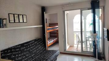 Una habitación pequeña y bien iluminada con sofá, literas, puerta corrediza de vidrio que da a un área exterior y un ventilador.