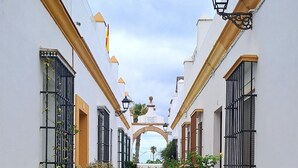 Uma estreita passagem pavimentada, ladeada por edifícios brancos com varandas, plantas em vasos e um céu claro.