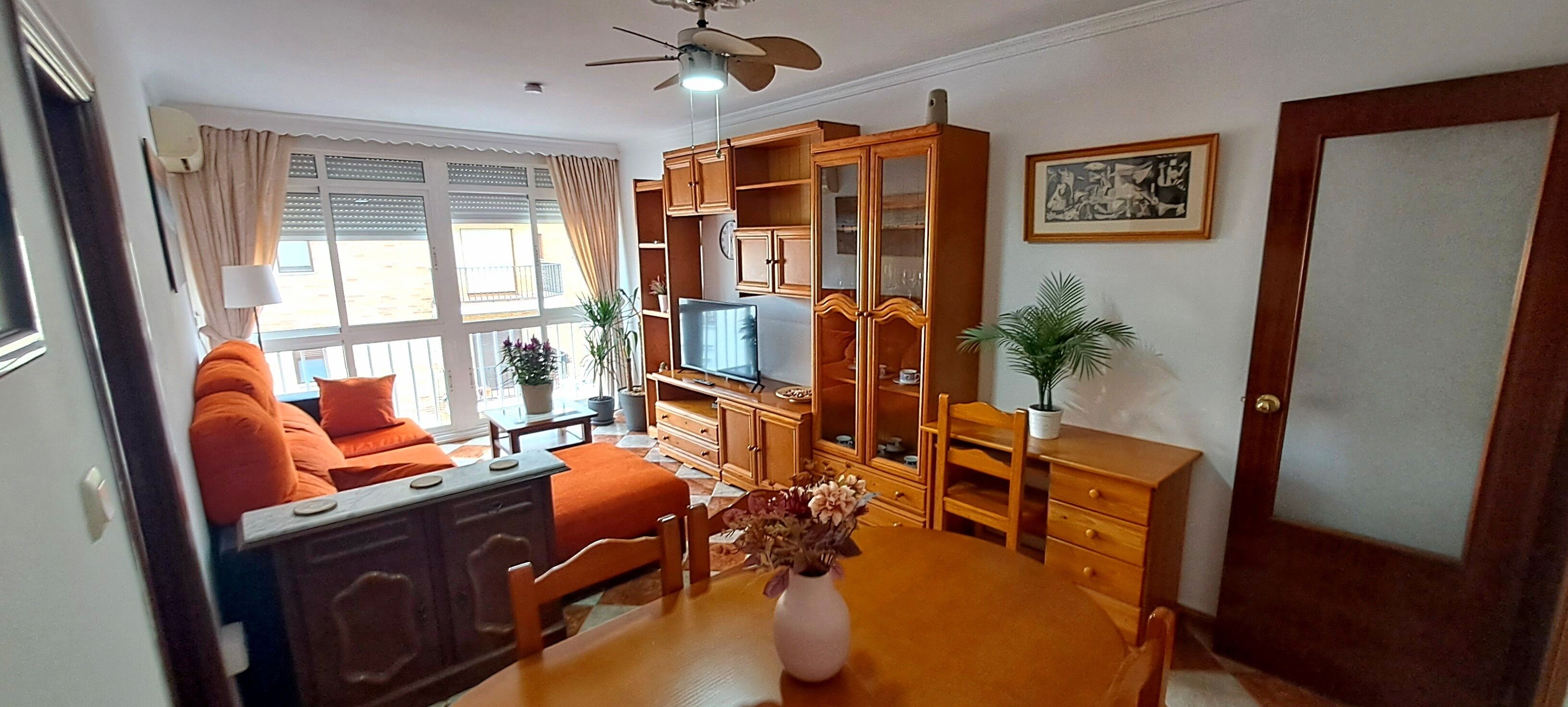 Sala de estar con sofá, televisión, armarios de madera y una mesa de comedor con un jarrón de flores.