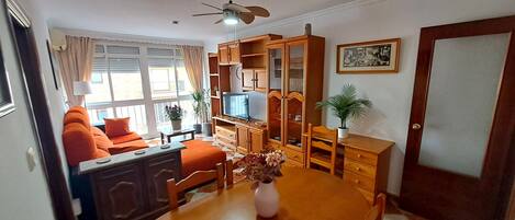 Sala de estar con sofá, televisión, armarios de madera y una mesa de comedor con un jarrón de flores.