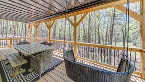 A covered outdoor deck with a table and chairs, surrounded by a wooden railing with a view of a forest. - Catalytic Derby themed (Helen)