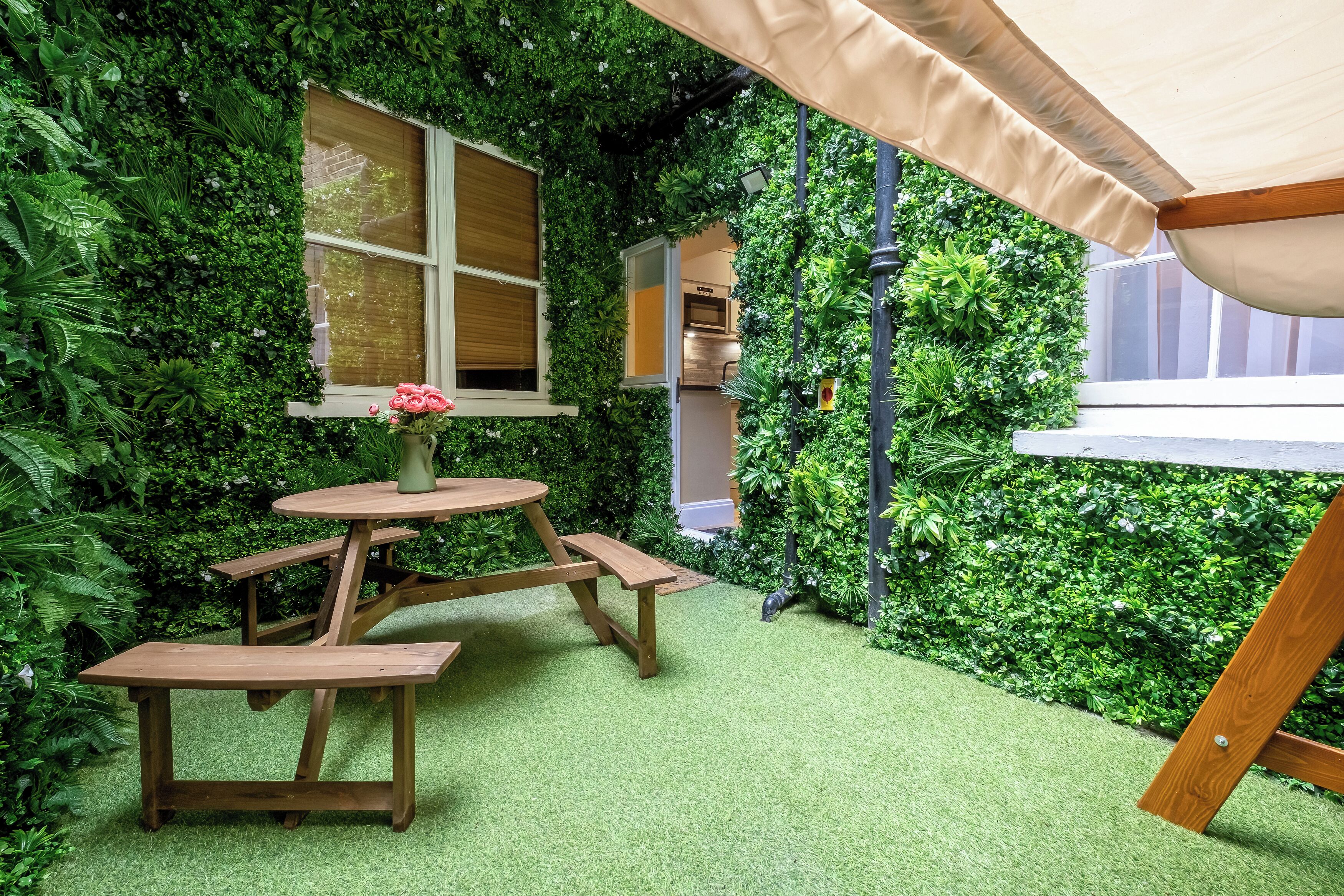 A cozy outdoor seating area with a wooden table and benches, surrounded by lush greenery and a small window.