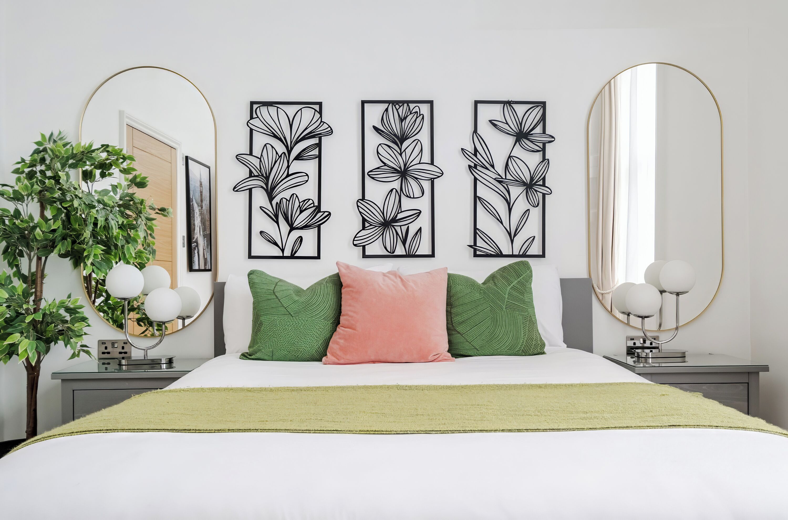 Ein Schlafzimmer mit einem Bett, grünen und rosafarbenen Kissen, einer grünen Tagesdecke, einer Pflanze und drei gerahmten Blumenbildern an der Wand.
