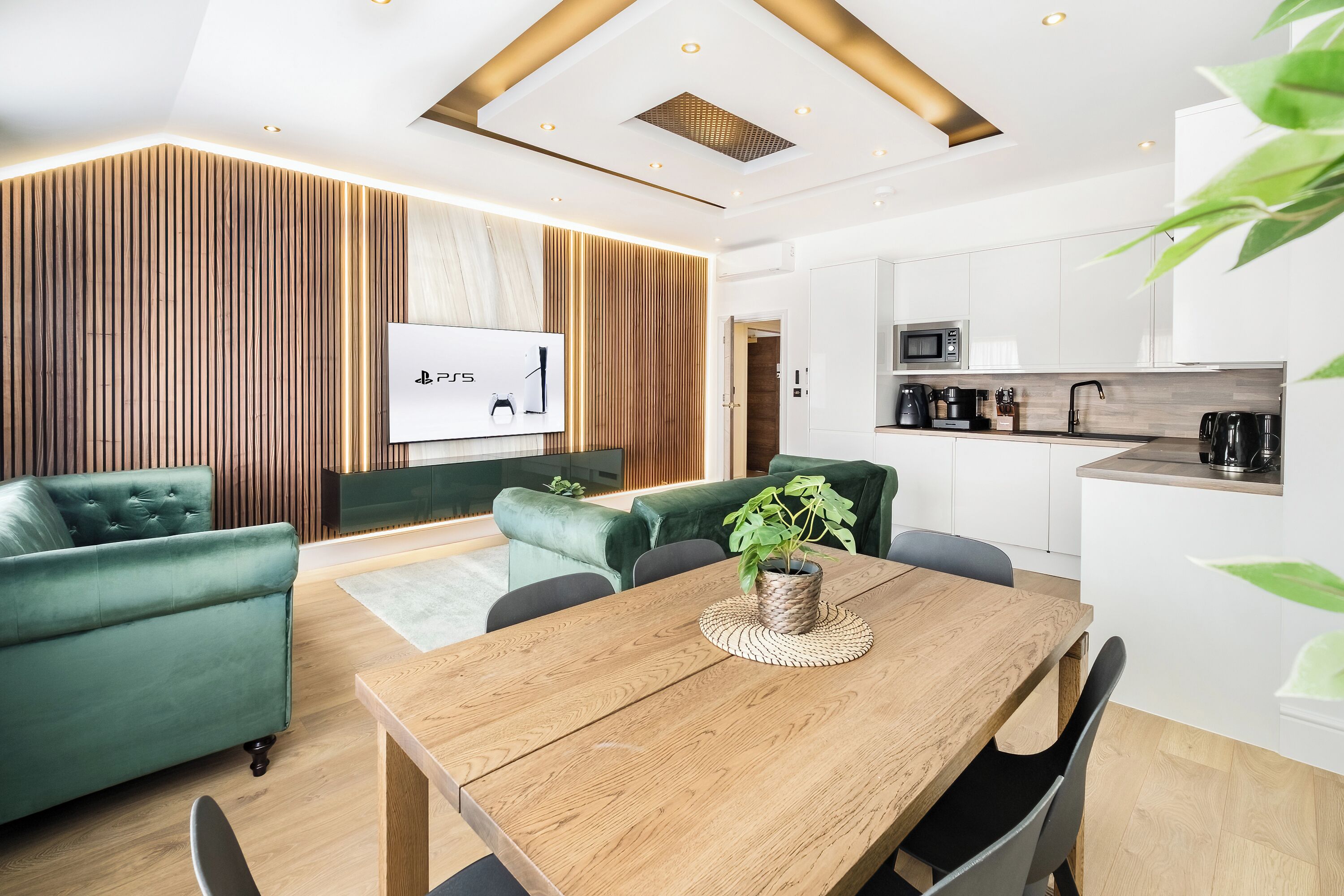 Ein modernes Wohnzimmer mit einem Holzespeisetisch, grünen Sofas und einer Küchenzeile mit weißen Schränken und Geräten.