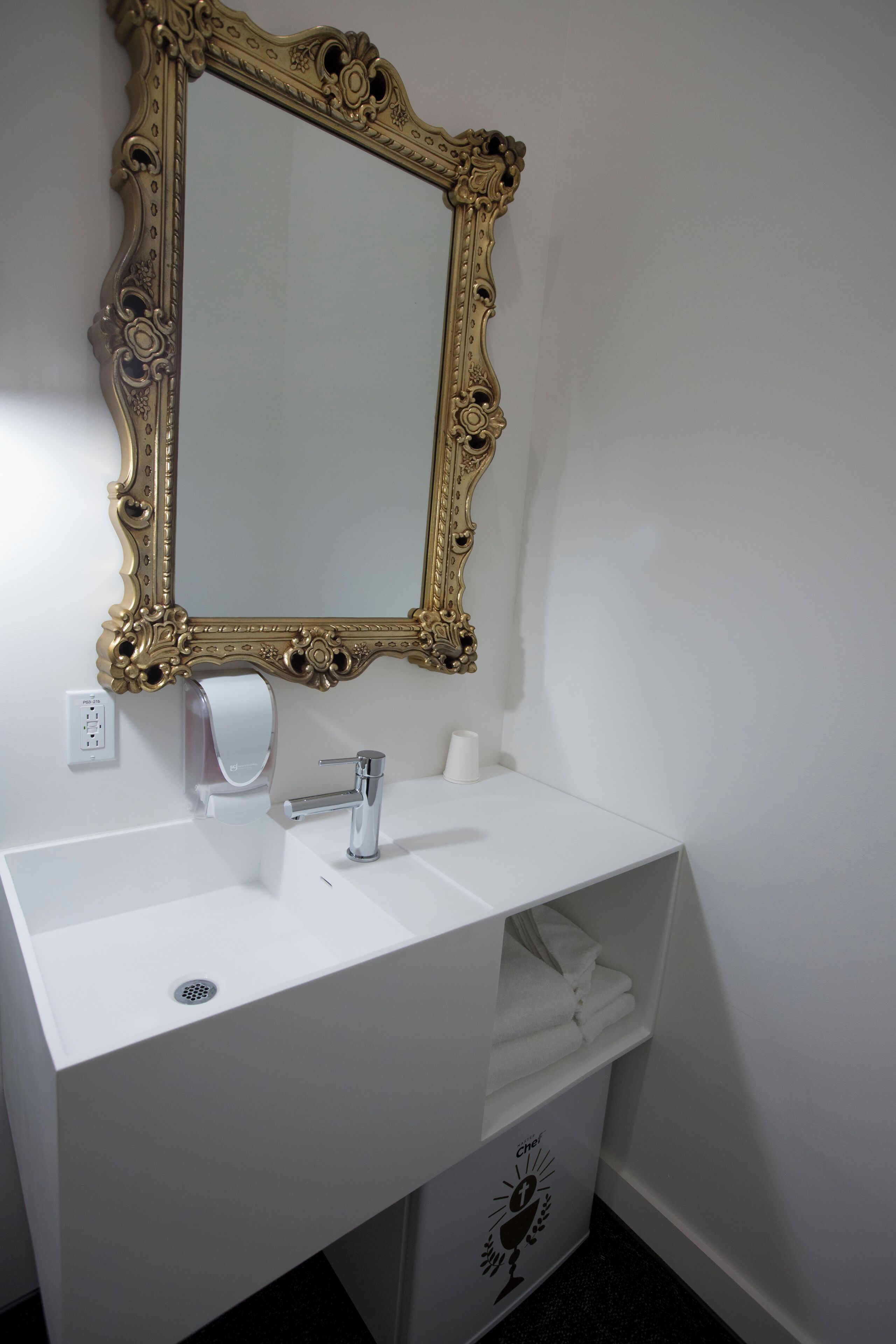 A bathroom with a white sink, a gold-framed mirror, a paper towel dispenser, and a towel rack.