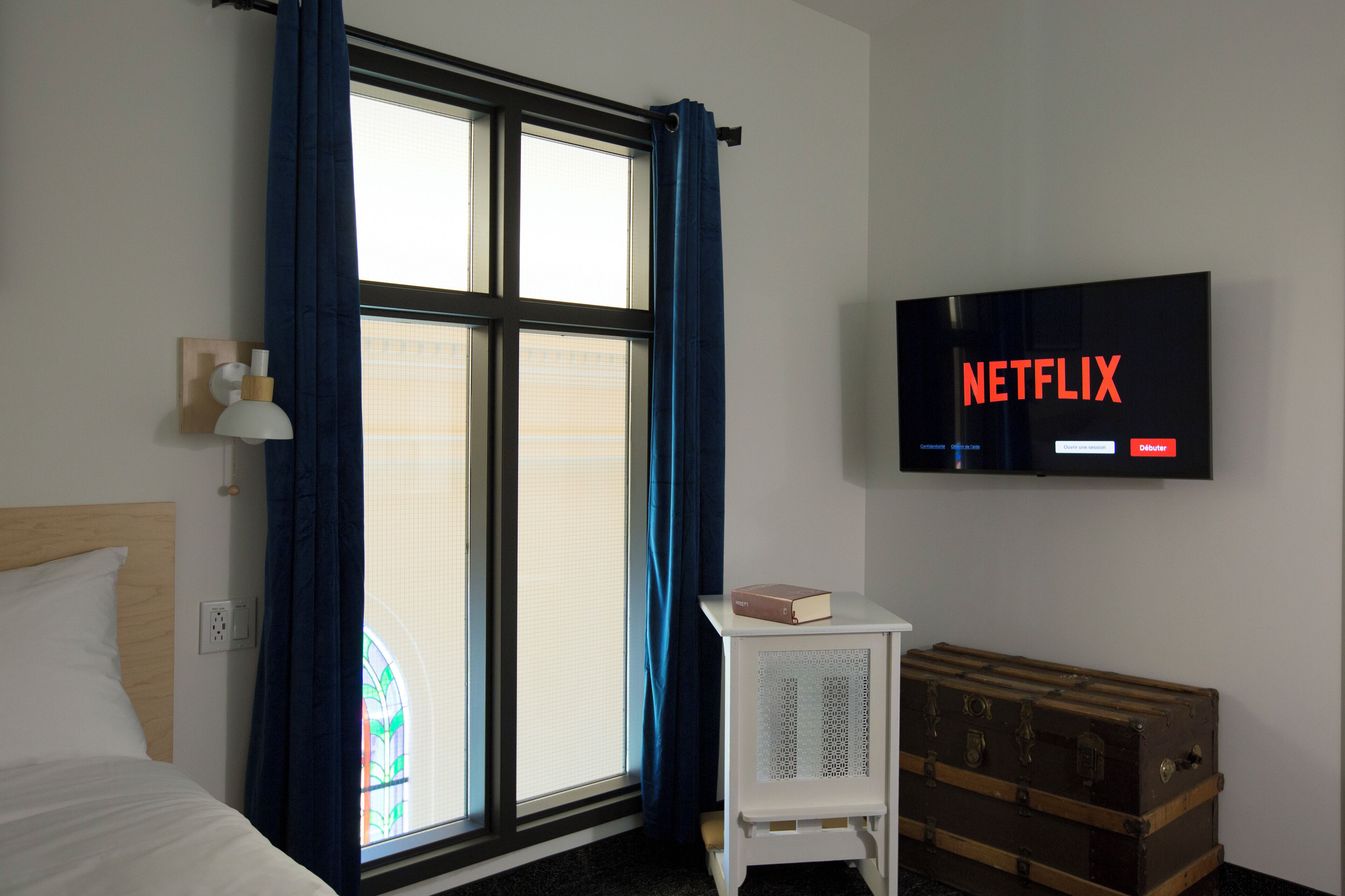 Habitación de hotel con cama, ventana con cortinas, un televisor mostrando 'NETFLIX', una mesita con un libro y un cofre de madera.