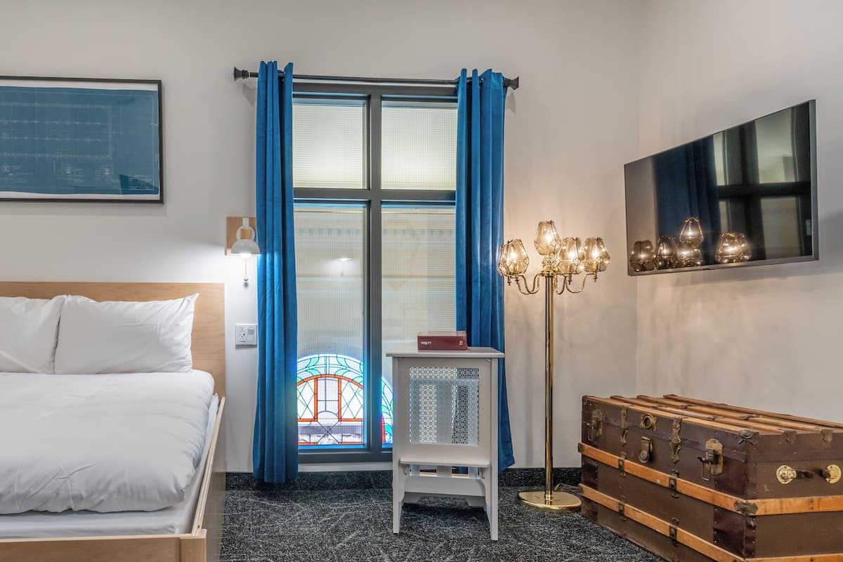 A hotel room with a bed, a small table with a lamp, a window with blue curtains, and a wall-mounted TV.
