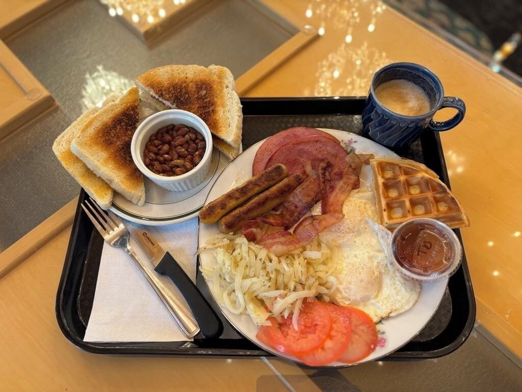 A breakfast tray with toast, beans, bacon, sausages, scrambled eggs, tomatoes, waffles, and a cup of coffee.