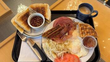 A breakfast tray with toast, beans, bacon, sausages, scrambled eggs, tomatoes, waffles, and a cup of coffee.