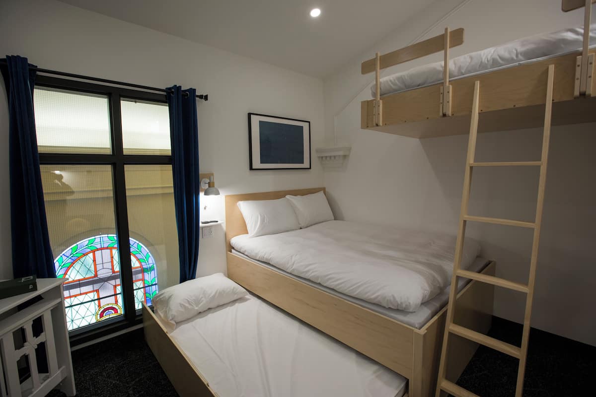A room with a bunk bed, a window with curtains, a bedside table, and a framed picture on the wall.