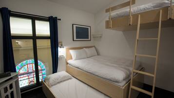 A room with a bunk bed, a window with curtains, a bedside table, and a framed picture on the wall.