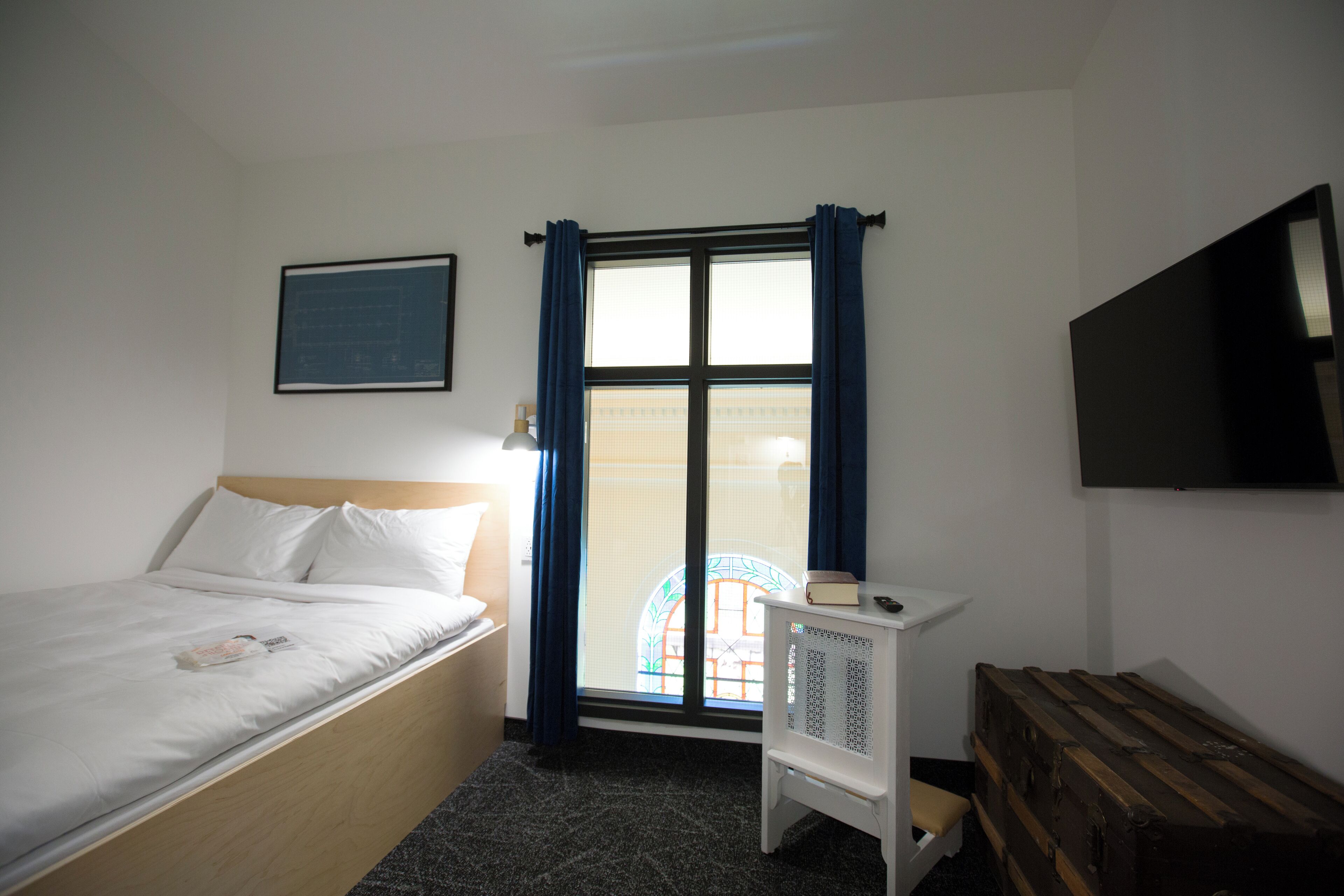 A bedroom with a bed, a window with curtains, a radiator, a television on the wall, and a wooden bench.