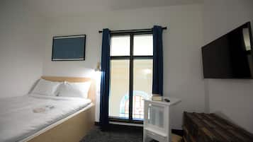 A bedroom with a bed, a window with curtains, a radiator, a television on the wall, and a wooden bench.