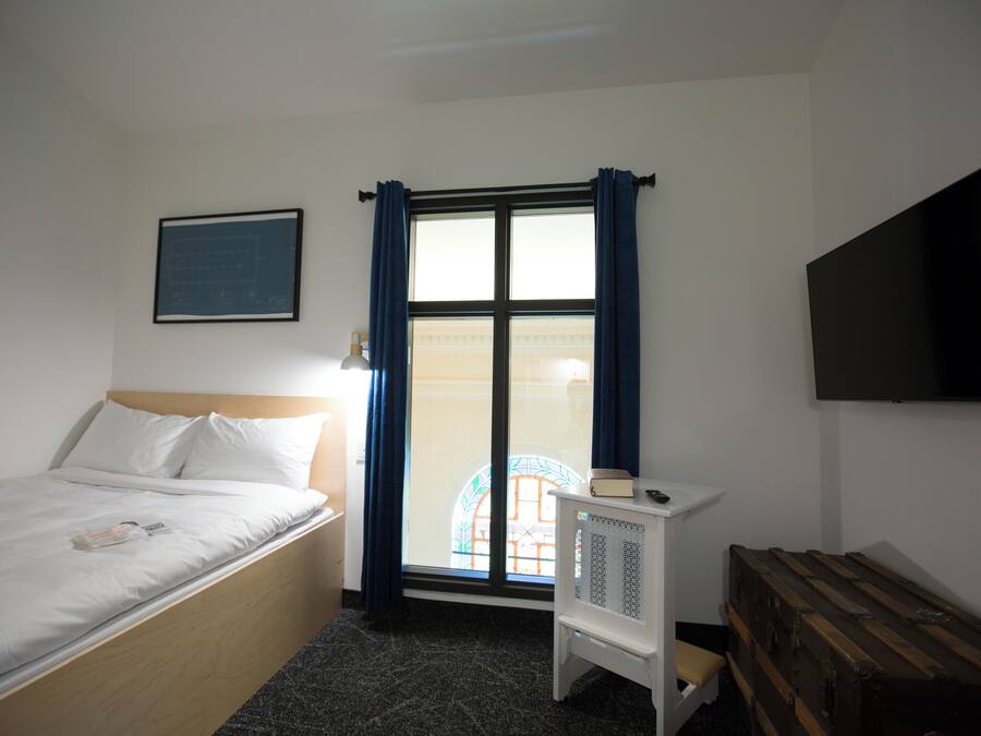 A bedroom with a bed, a window with curtains, a radiator, a television on the wall, and a wooden bench.