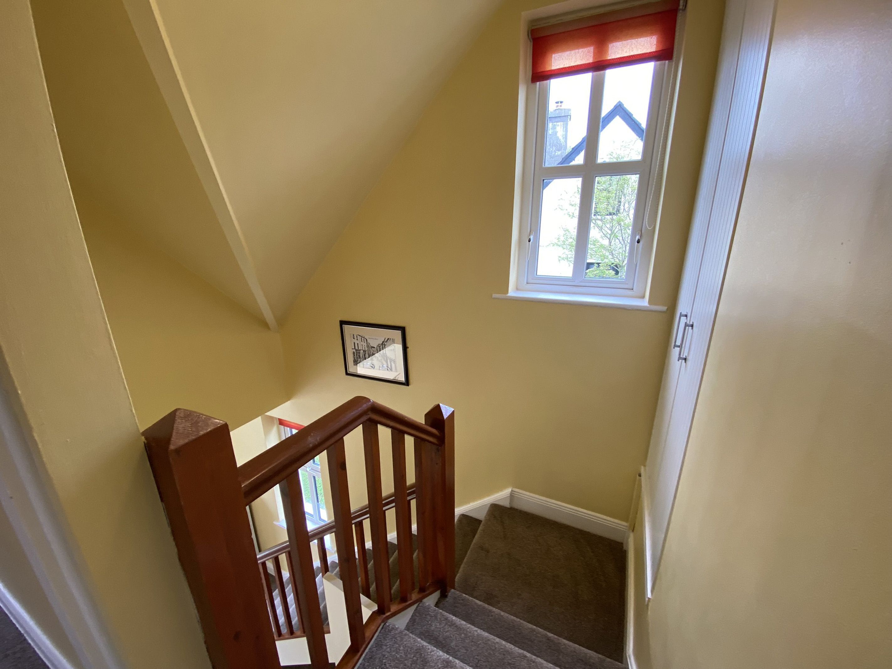 A staircase with wooden railings leading to a landing. A window with a red valance is visible, and a framed picture hangs on the wall.