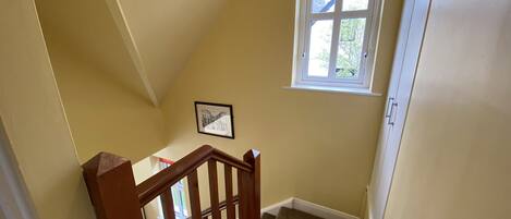 A staircase with wooden railings leading to a landing. A window with a red valance is visible, and a framed picture hangs on the wall.