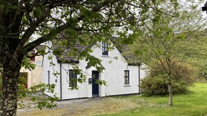 A white building with a dark blue door and windows, surrounded by a grassy area and trees. - Cottage 466 - Clifden - sleeps 5 guests  in 3 bedrooms (Clifden)
