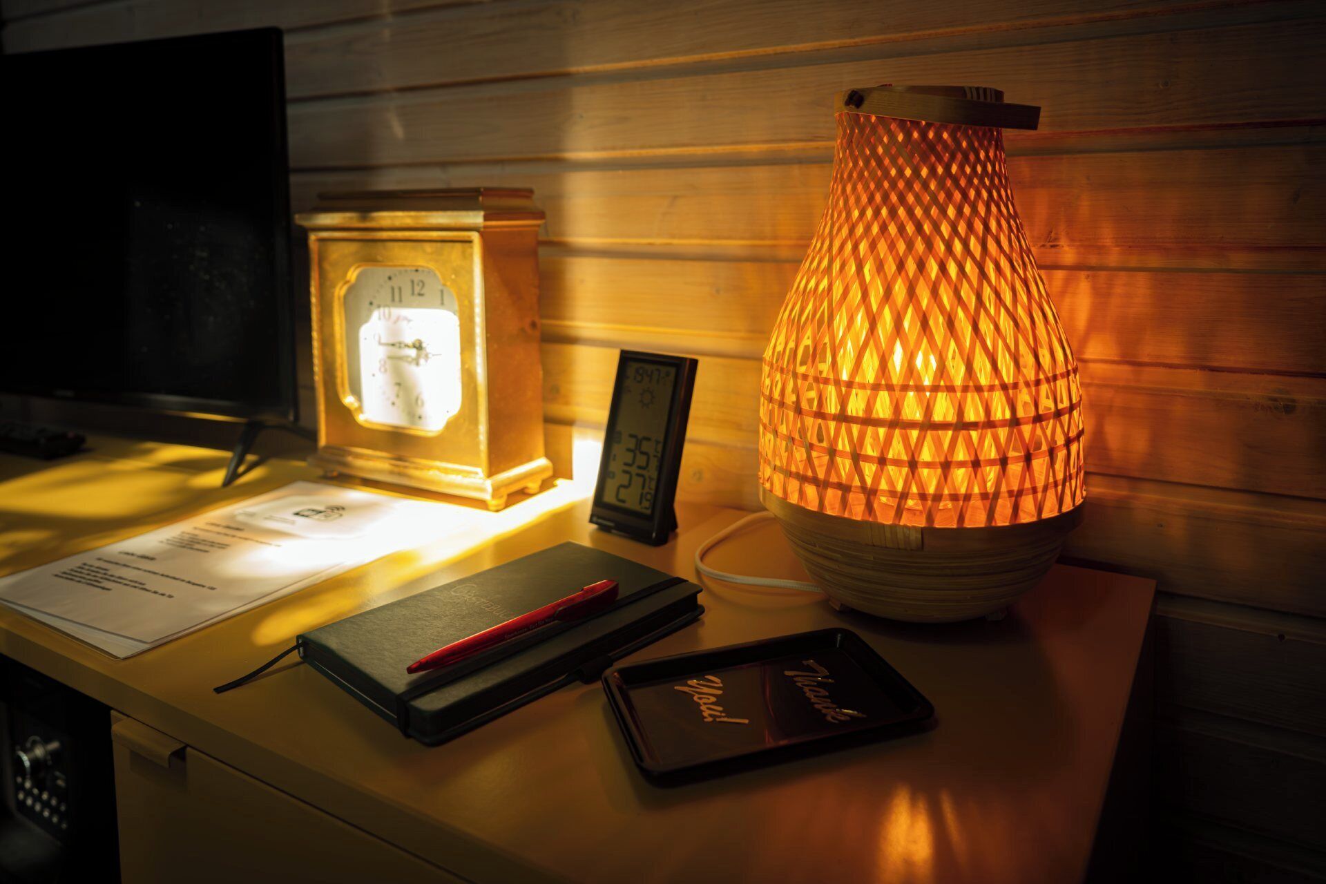 A wooden table with a vintage clock, a modern digital thermometer, a decorative lamp, and a notebook with a red pen.