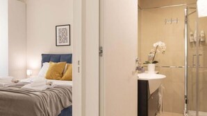 Quarto de hotel com cama, mesa de cabeceira, banheiro com chuveiro e pia.