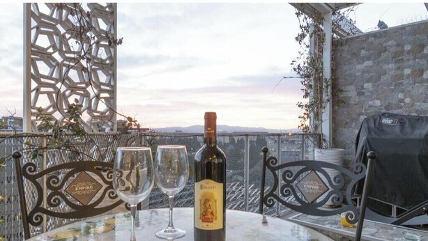 Uma mesa com uma garrafa de vinho e duas taças, com vista para uma paisagem urbana e uma cerca metálica decorativa.