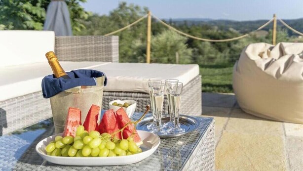 Een tafel met een champagnefles, watermeloenpartjes en druiven, op een terras met uitzicht over het landschap.