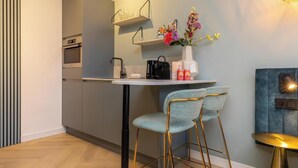 A modern kitchen with a central island, a built-in oven, and a wall-mounted shelf with glassware. There are two upholstered chairs with gold legs and a small round table with a gold base. - Studio - Van der Manderelaan 16 'Studio B' | Koudekerke (Koudekerke)