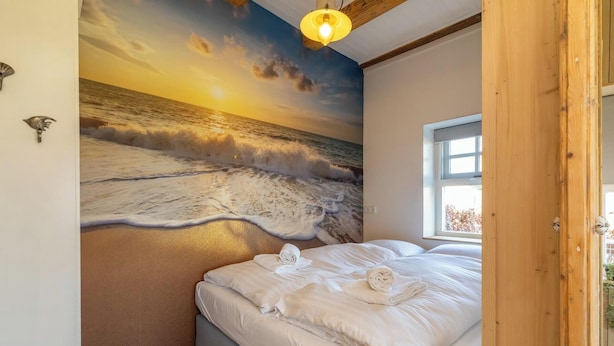 Un dormitorio con un mural de playa al atardecer, una cama con ropa blanca y una ventana con vistas al exterior.