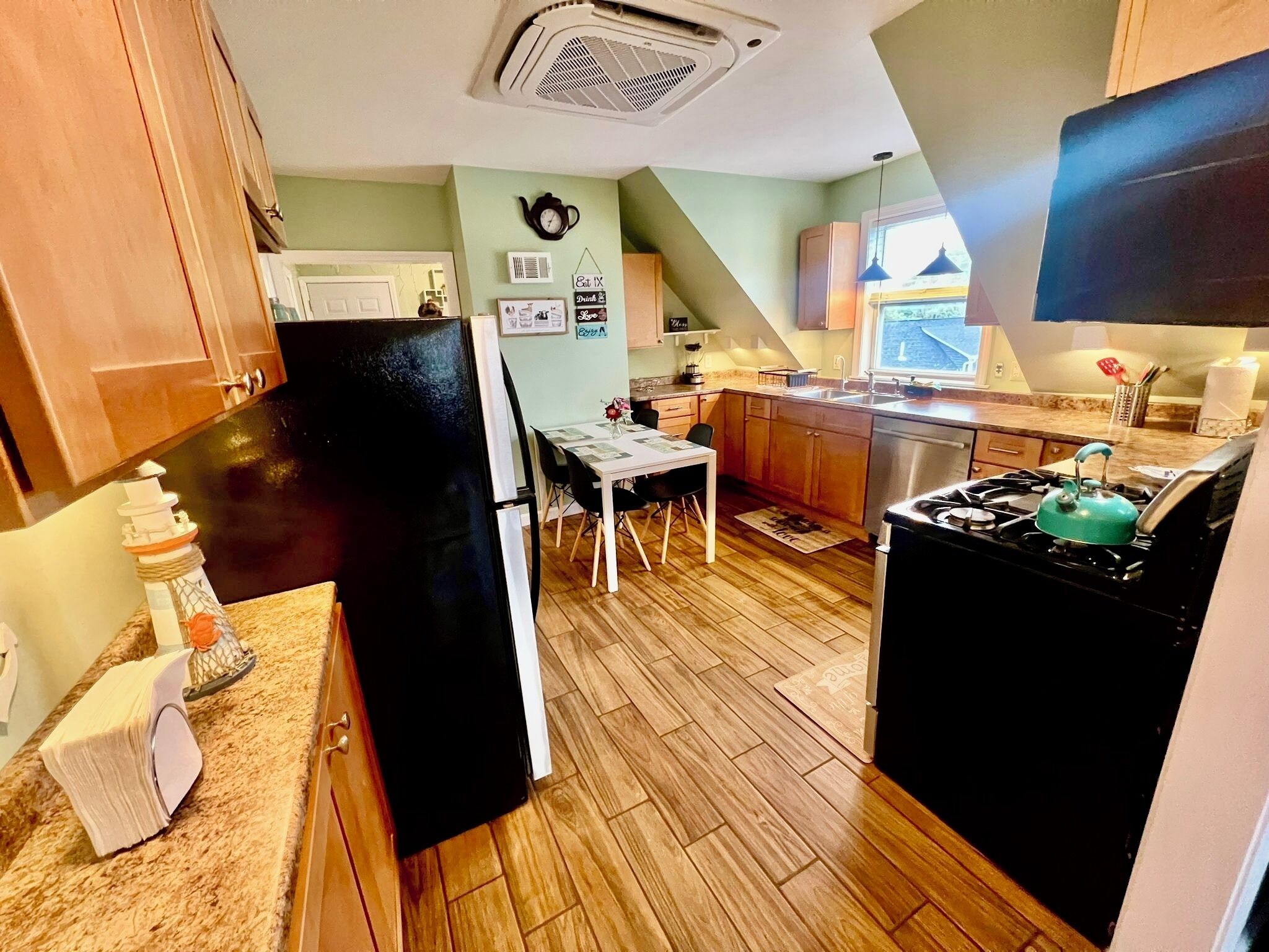Una cocina compacta con gabinetes de madera, un refrigerador negro, una mesa de comedor con sillas, una estufa a gas y un ventanal.