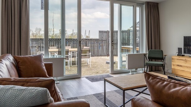 Un salon avec un canapé en cuir, une table basse en bois et une vue, grâce à de grandes portes vitrées, sur une terrasse extérieure dotée d’une table et de chaises en bois.