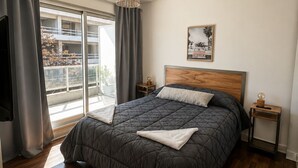A bedroom with a large bed, a wooden headboard, a bedside table with a lamp, a window with curtains, and a view of buildings outside. - GURRUCHAGA PALERMO (Buenos Aires)