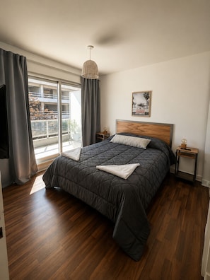 Un dormitorio con una cama grande, un cabecero de madera, una mesita de noche con una lámpara, una ventana con cortinas y vistas a edificios.