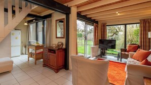 Ein großzügiger Wohnbereich mit Sofa, Fernseher und Esstisch. Eine Treppe führt auf eine obere Ebene, und eine Glasschiebetür öffnet sich zum Garten.