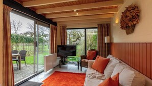 Ein Wohnzimmer mit einem weißen Sofa, orangefarbenen Kissen, einem Fernseher und einem großen Fenster mit Blick auf einen Garten.