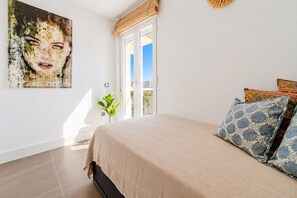 A bedroom with a bed, a window, a plant, and a painting on the wall.