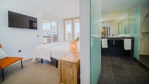 Una habitación de hotel moderna con una cama, una mesita de noche de madera, un televisor y un baño con cabina de ducha de vidrio.