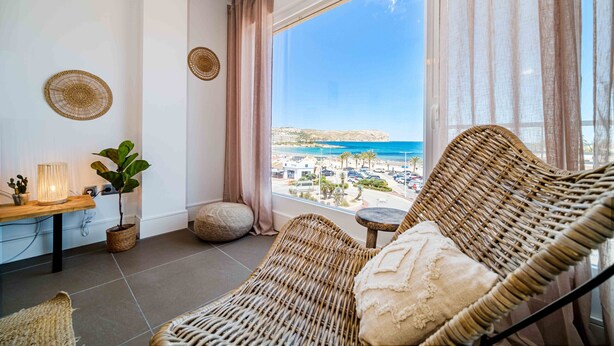Un balcón con una silla de mimbre, una mesita con una lámpara, una planta en maceta y vistas al mar y a edificios.