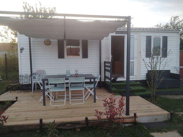 Een overdekt terras met een tafel en stoelen, een klein tuin en een witte stacaravan met luiken.