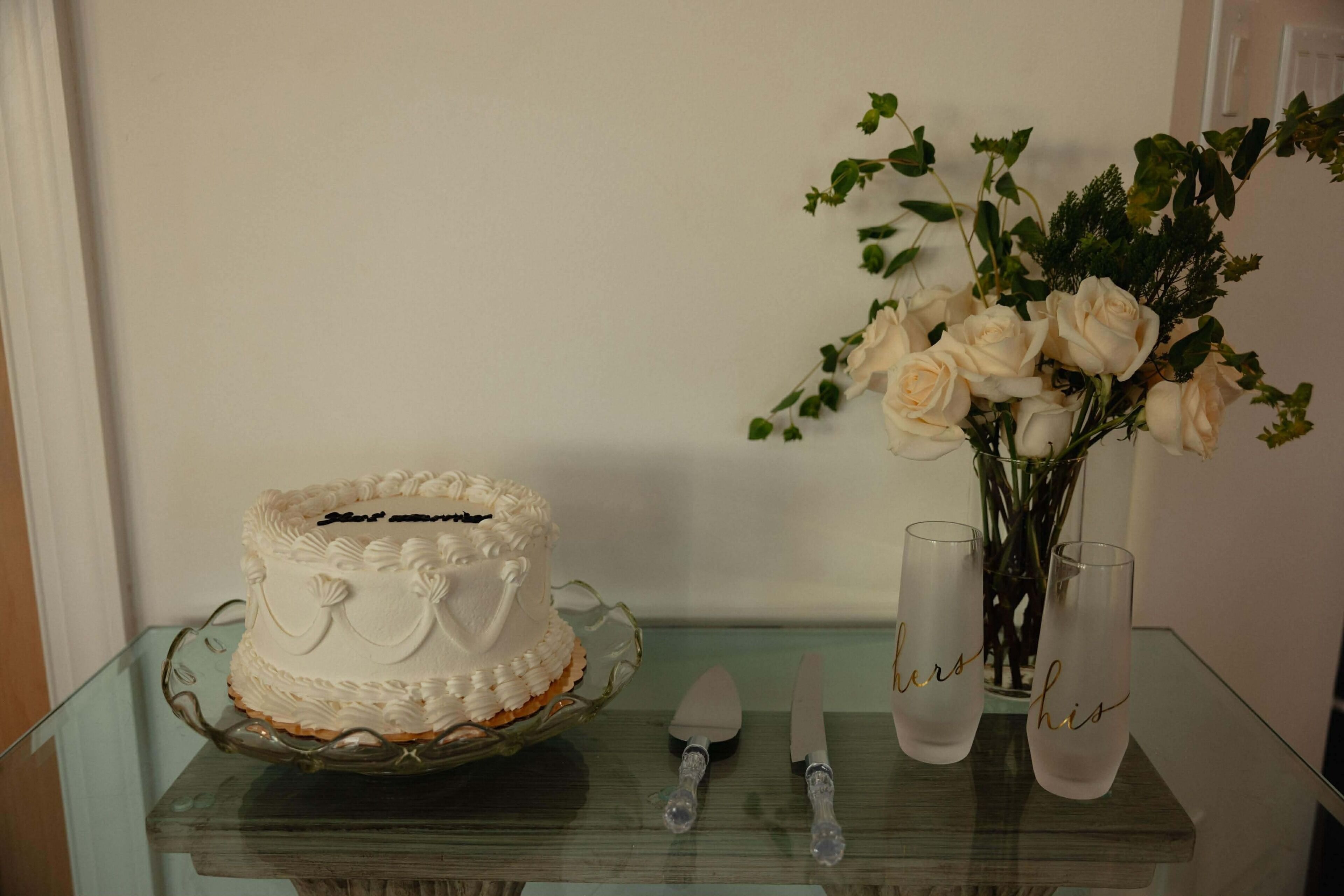 Un gâteau sur une table en verre, avec un vase de roses blanches et deux verres sur lesquels sont inscrits les mots « à elle » et « à lui ».