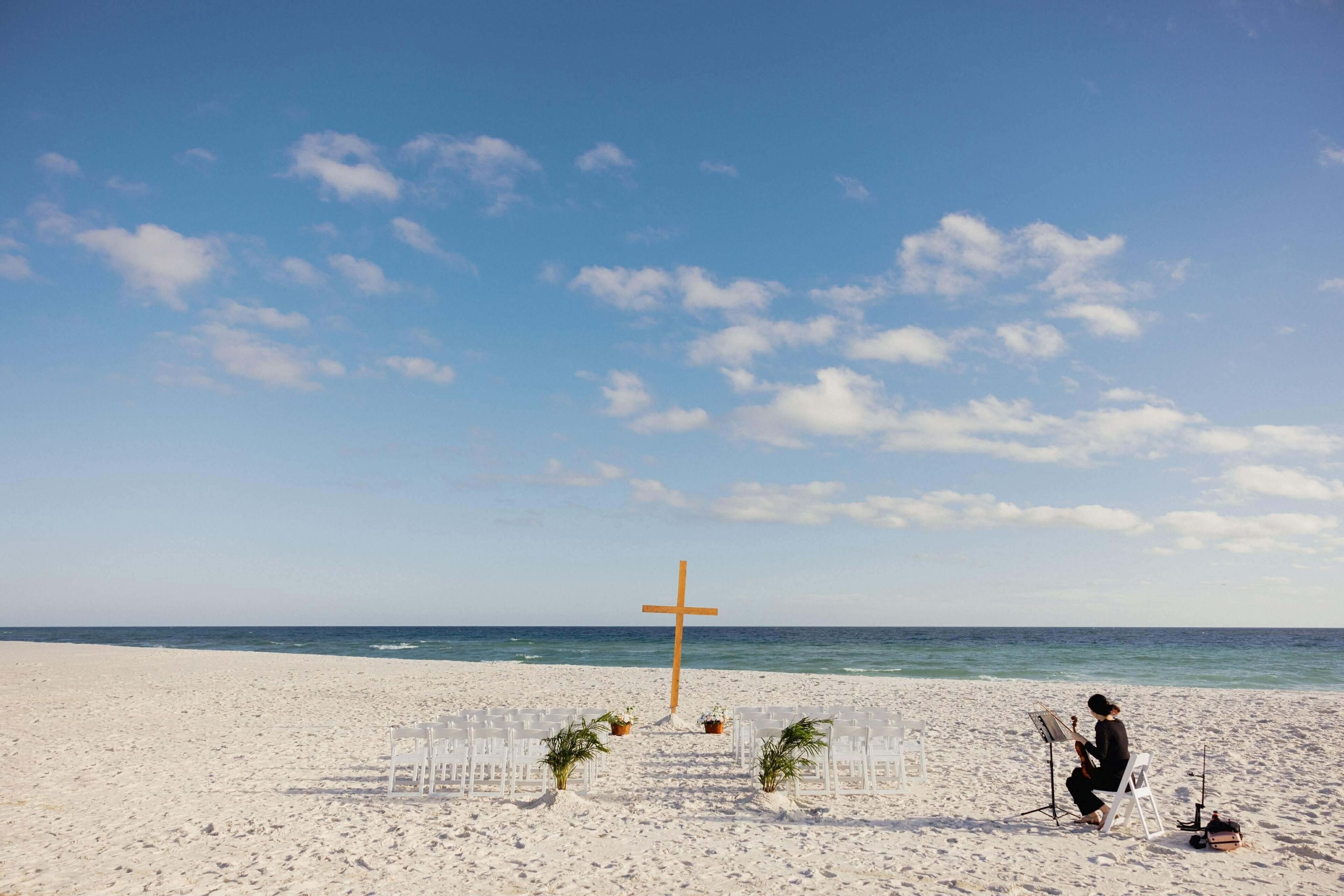 A beach setting with a wooden cross, white chairs, and a person playing a guitar.