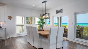 A dining area with a wooden table and white chairs, large windows with a view of the beach, and a ceiling light fixture. - Beachfront Luxury Estate Sleeps 20 & Allows Events (Navarre)