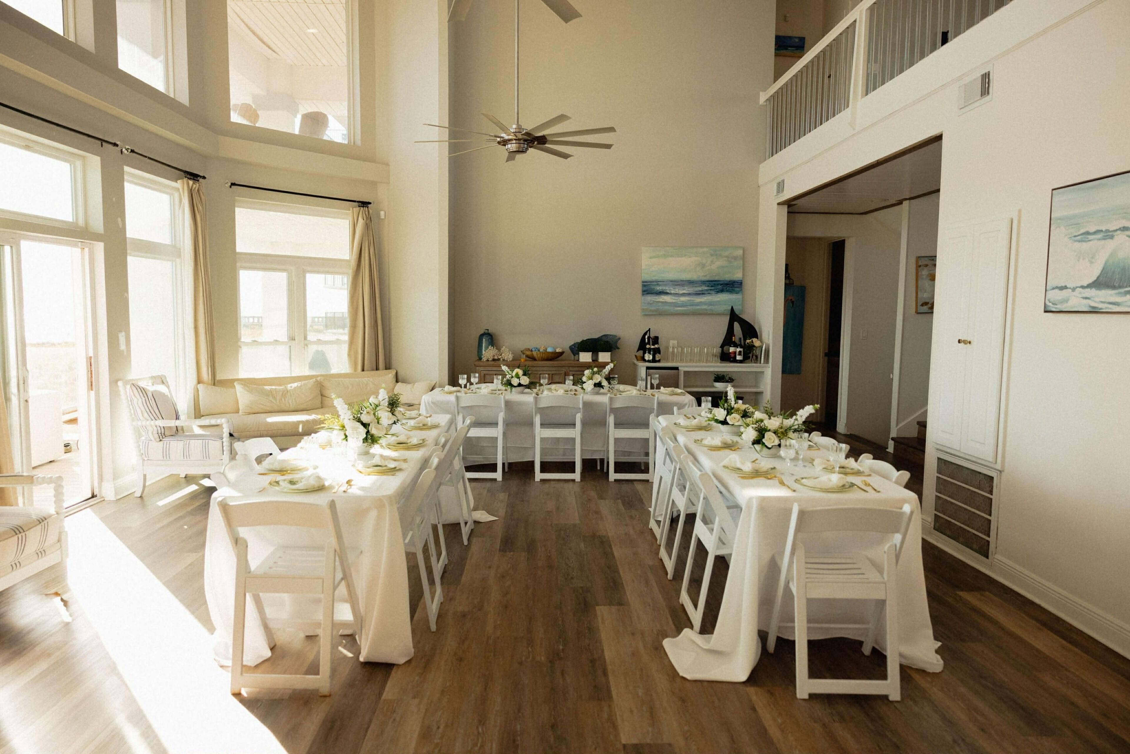 Un espace repas avec plusieurs tables dressées pour le repas, un ventilateur de plafond et une vue dégagée sur l’extérieur grâce à de grandes fenêtres.