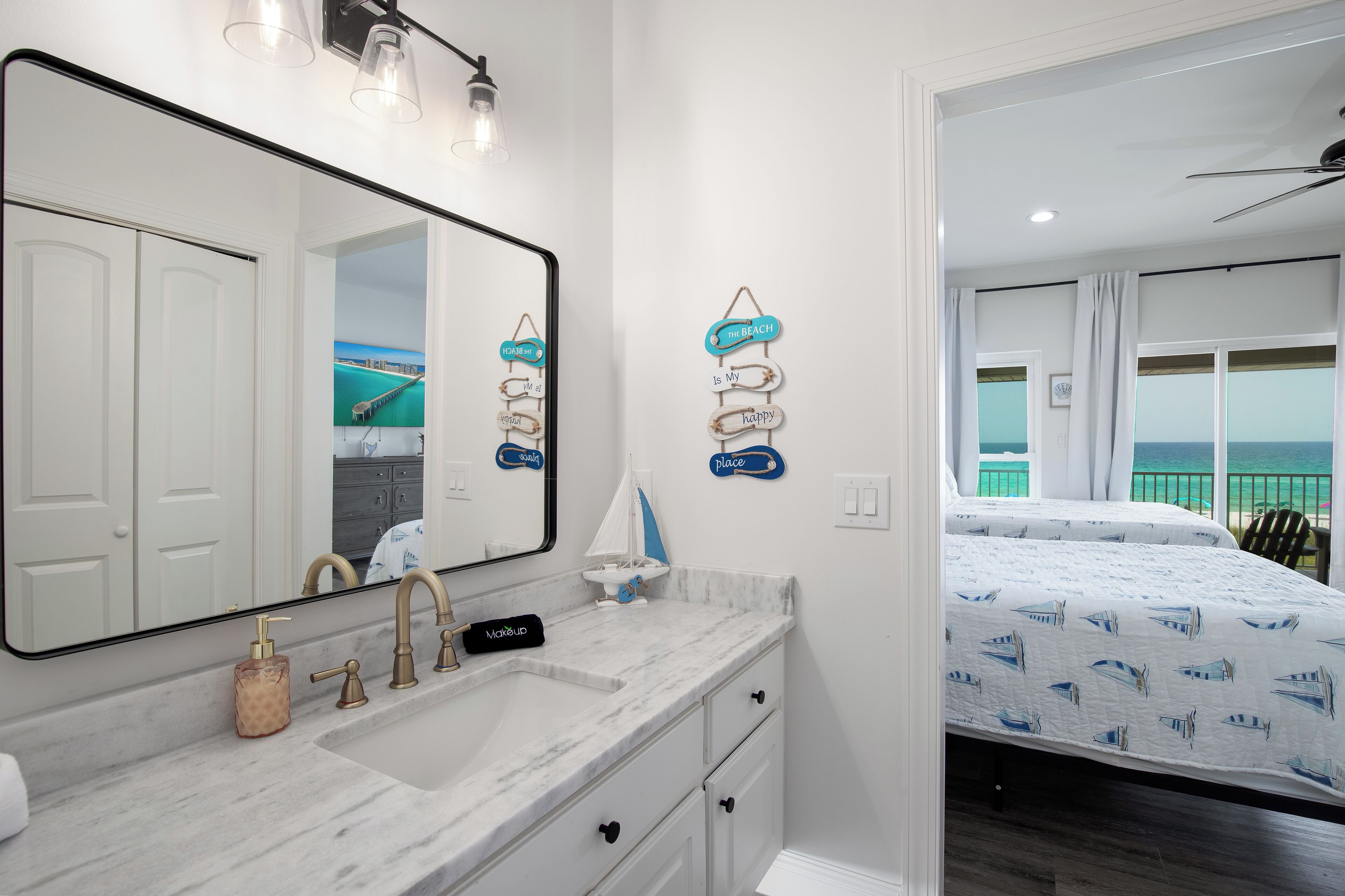 A bathroom with a large mirror, a sink with gold faucets, a soap dispenser, and a towel. There is a bed with a blue and white patterned bedspread in the adjacent room.