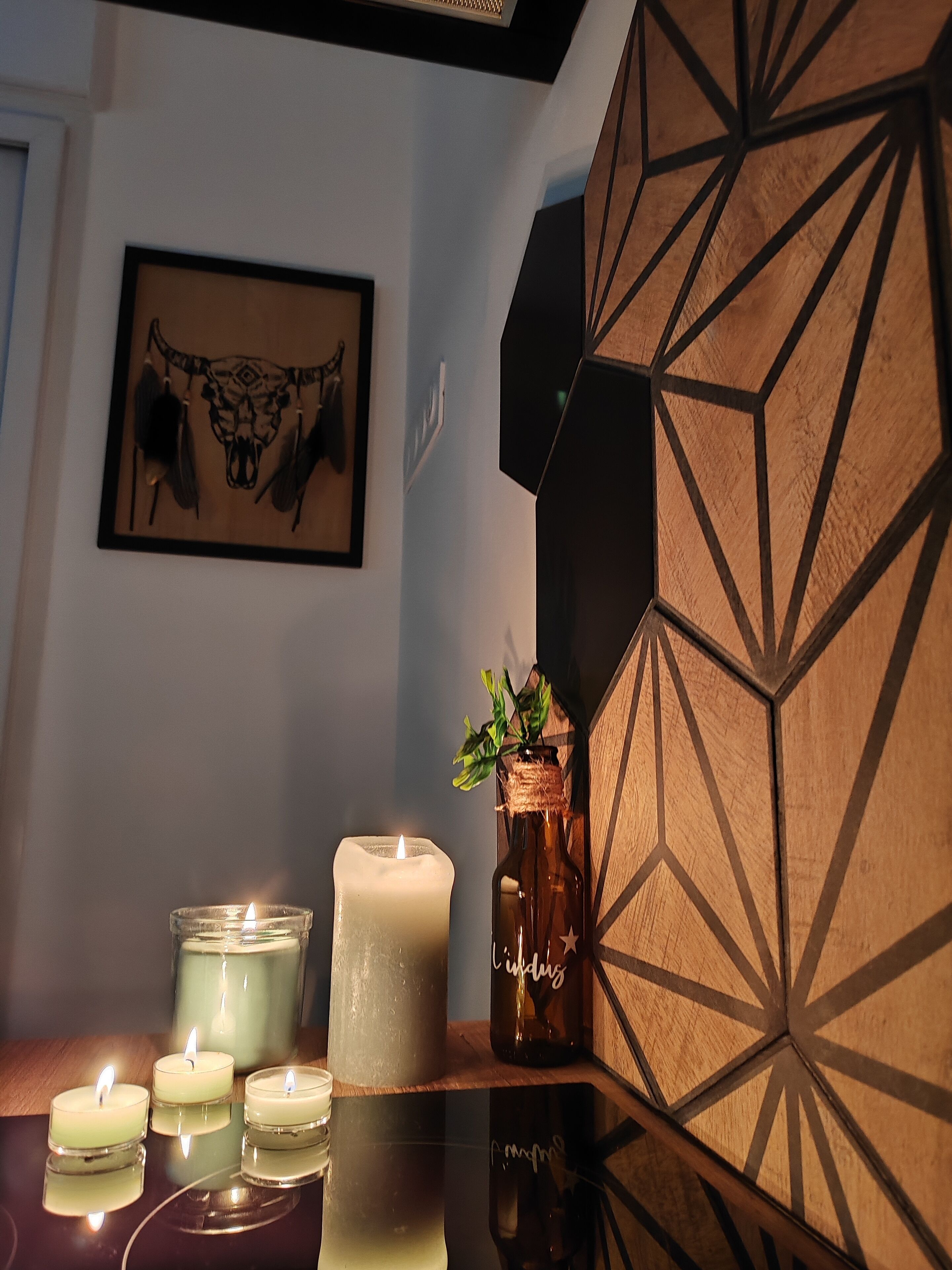 Una stanza con un muro a motivi geometrici, un quadro incorniciato e diverse candele accese su una superficie riflettente.